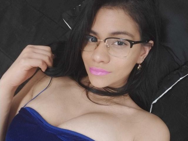 AnastasiiaS with black hair, glasses, pink lipstick, blue velvet top, showing her tits, lying on a black surface, hand in hair, looking at camera, earrings visible.