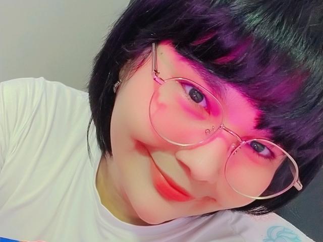 PrittyAlicia with pink-tinted glasses, black bob haircut, and white shirt, smiling with red lips, showing a cute, chubby face. Her tits are small and barely visible.