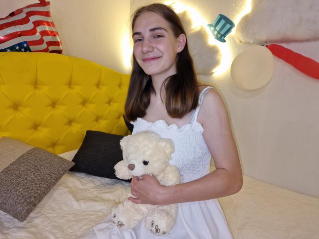 NikaStark with brown hair, smiling, wearing a white dress, holding a fluffy white teddy bear, sitting on a yellow tufted bed with American flags.
