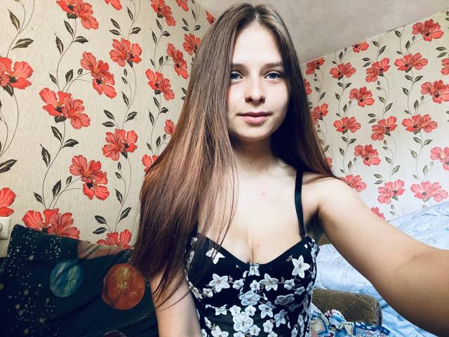 RoxannaMary with long brown hair, blue eyes, and small tits in a black floral dress, taking a selfie in a room with red flower wallpaper.