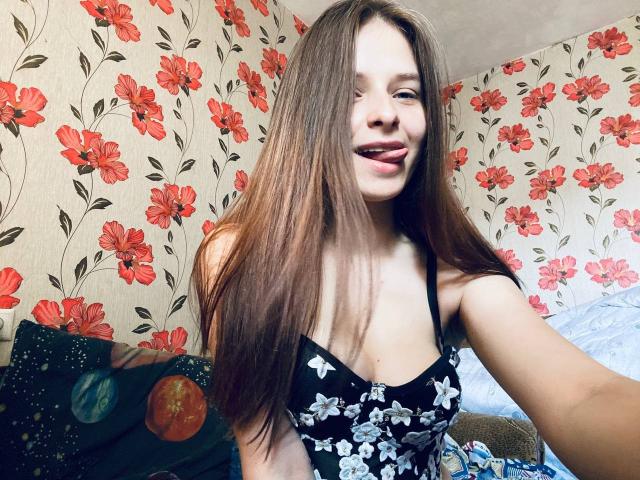 RoxannaMary with long brown hair, smiling, wearing a black floral dress, showing her small tits, in a room with red floral wallpaper, taking a selfie.