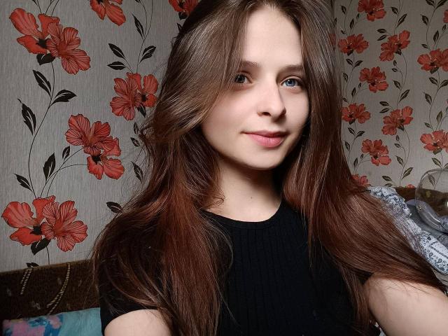 RoxannaMary with long brown hair, blue eyes, and a slight smile, wearing a black ribbed shirt, showing her cute face and natural tits in a casual selfie.