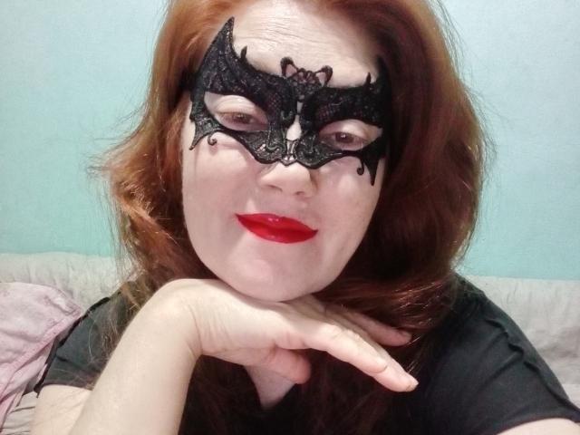 SadieLorri with red lipstick and black lace mask, resting chin on hand, has pale skin, red hair, and is wearing a black shirt. Her mask has ornate, dark patterns.