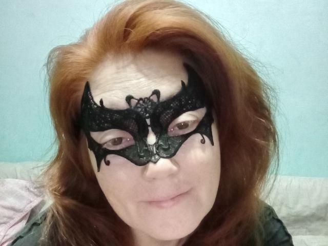 SadieLorri with red hair, wearing a black lace mask, smiling slightly, showing pale skin and brown eyes, wearing a dark green top.