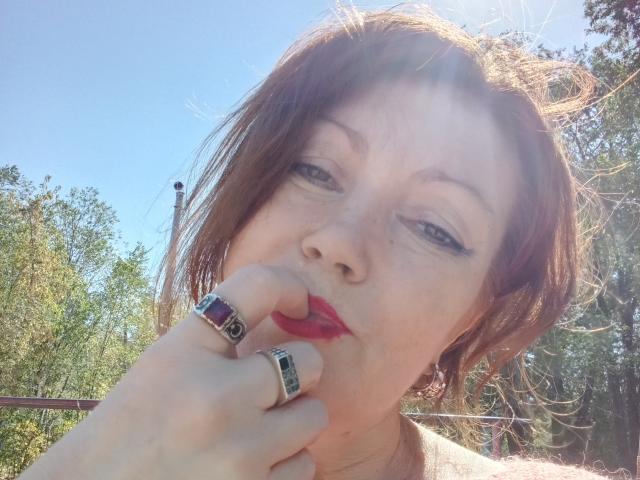 SadieLorri with short brown hair, blue eyes, and red lipstick, biting her finger with a ring on her hand, sunlight in her face, wearing a silver ring.