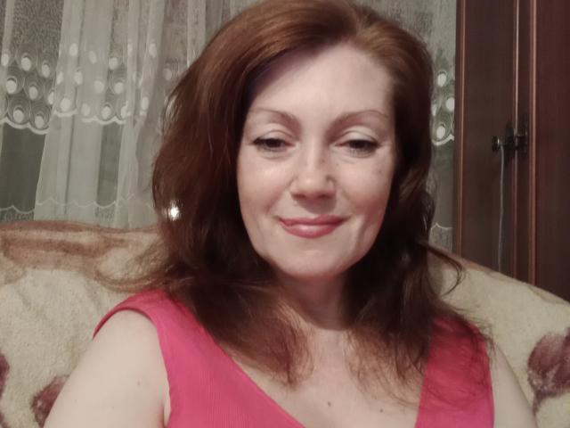 SadieLorri with red hair, smiling, wearing a pink top, showing her tits, fair skin, subtle makeup, and small earrings, sitting on a beige patterned couch.