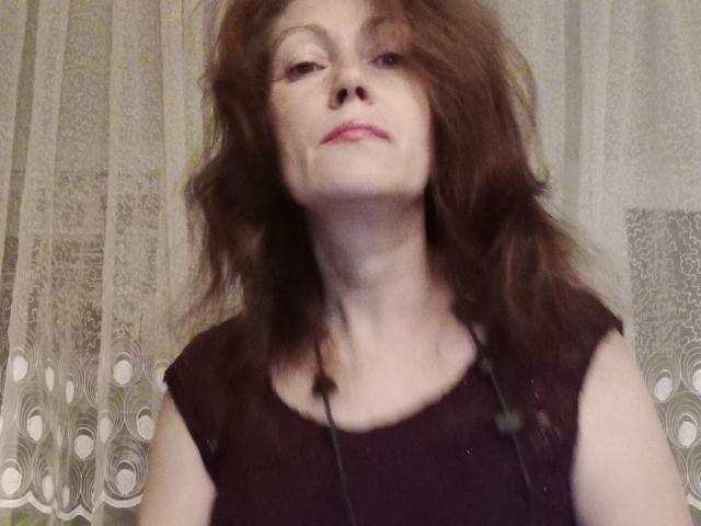 SadieLorri has messy brown hair, a pale face, and wears a black sleeveless top with a necklace. She looks up with a slightly bored or indifferent expression.