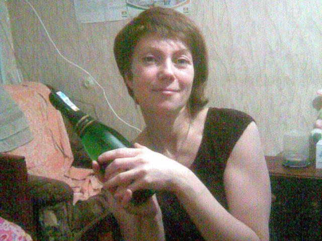 SadieLorri with short brown hair, black sleeveless top, holding a green bottle in both hands, smiling slightly, in a dimly lit room. Her tits are modestly covered.