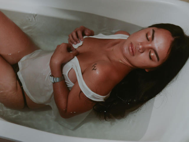 TransKatie in a white, wet, see-through tank top, lying in a bathtub with water, covering her tits with her hands, eyes closed, long dark hair, tattoo on right shoulder.