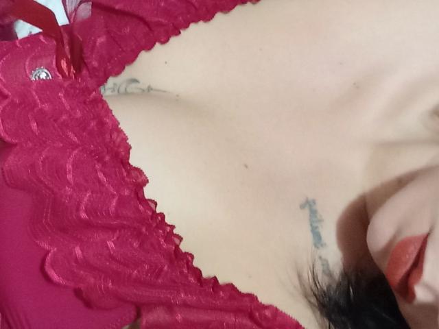 BiaSexy69's cleavage in a red lace bra, showing small tattoos on both sides of her chest, with her dark lipstick and black hair visible on the right.