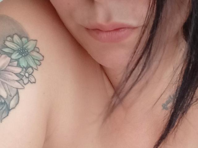 BiaSexy69's upper body, showing a tattoo of colorful flowers on her shoulder, with her lips slightly closed and black hair partially covering her face. Her tits are partially visible.