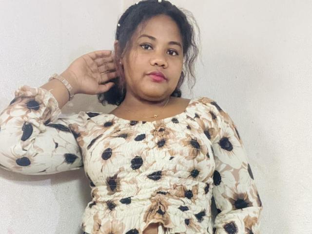 Candilla, a chubby woman with medium brown skin, wearing a floral top, has her hand on her head, looking at the camera with pink lipstick.