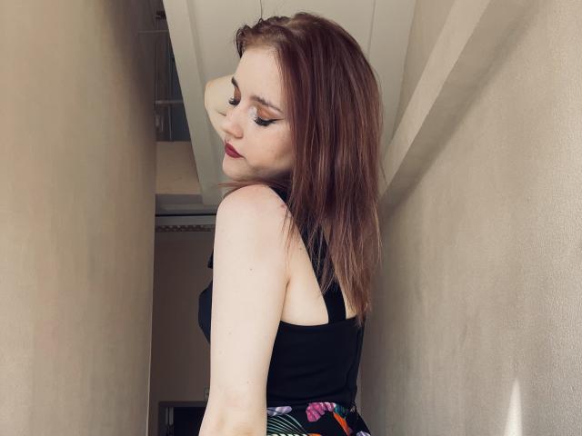 DaMa69s, avec sa peau pâle, ses cheveux roux-brun et son rouge à lèvres foncé, portant un haut noir, regardant par-dessus son épaule, montrant ses petites seins et ses fesses, dans un couloir étroit..