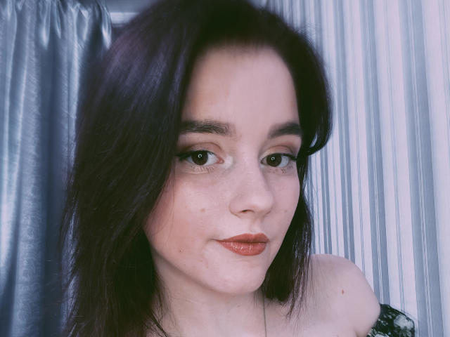 DaMa69s has pale skin, dark brown hair, green eyes, and red lipstick. She's looking at the camera with a slight smile, showing off her bare shoulders.