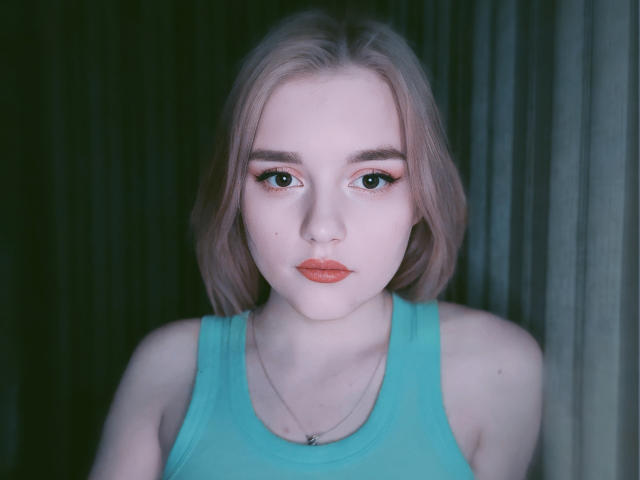 DaMa69s has pale skin, pinkish-blonde bob, green eyes, and wears a teal tank top with a delicate necklace, looking straight into the camera with a serious expression.