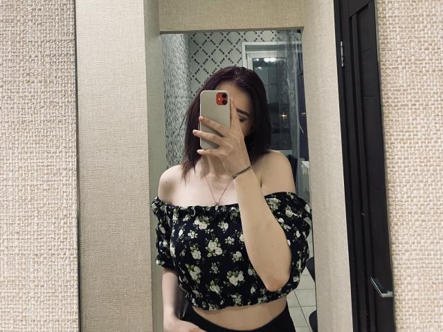 DaMa69s with medium tits, black floral off-shoulder top, holding phone in mirror selfie, dark brown hair, beige walls, black door, wearing black bottoms, light skin, subtle makeup.