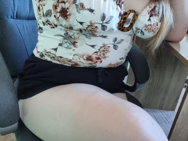 Leiliia sitting with her tits in a floral top, black shorts, and her chubby ass exposed. She's on a chair with a wooden desk to the right.