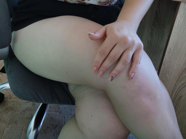 Leiliia's bleke, mollige dijen zijn gekruist, waarbij haar hand met roze nagels te zien is, rustend op haar rechter dij. Haar zwarte jurk is omhoog getrokken, waardoor haar billen zichtbaar zijn. Ze zit op een stoel..