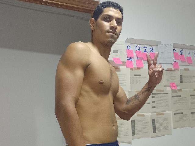 McBride, shirtless, standing, brown skin, short black hair, muscular, tattoos on right forearm, making a peace sign, wearing blue pants, in a room with white walls and notes.