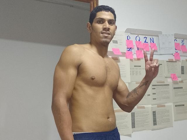 McBride, shirtless, muscular, tan skin, short black hair, small tattoo on right forearm, dark blue pants, making a peace sign, standing in a room with pink sticky notes.