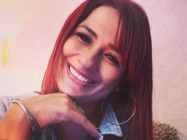 Amanda, com cabelo ruivo, sorrindo, segurando o queixo, usando brincos grandes de argola e uma camisa de jeans, mostrando dentes brancos e um nariz fofo. Sua mão está próxima ao rosto..