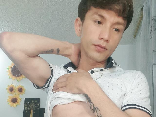 FlacoVergon69, brown-haired, lifts white polka-dot shirt, revealing flat chest and tattoos on arms, with a slight pout, in a well-lit room.