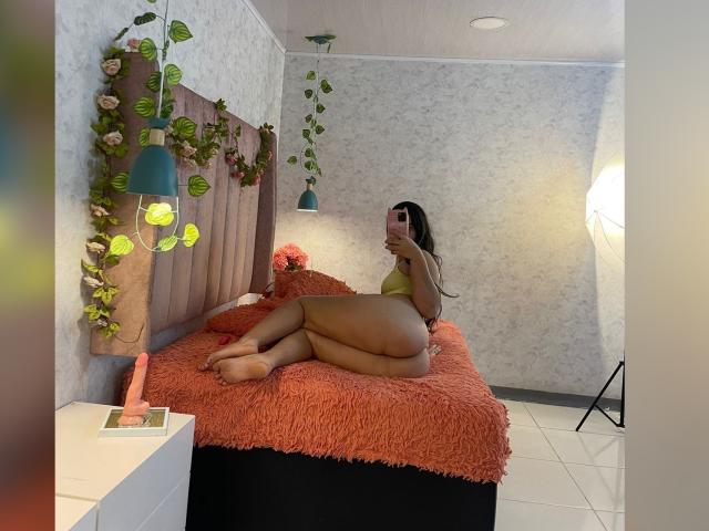 DanTaylor mit kurvigen Hüften, gebräunter Haut und langen braunen Haaren, liegt auf einem Bett mit einer orangefarbenen, flauschigen Decke, trägt ein grünes Oberteil und macht ein Selfie mit einem rosa Handy..