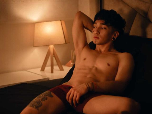 RossDiamond69 is a muscular, shirtless guy with tattoos, dark curly hair, and ear gauges, wearing red briefs, reclining on a bed with a lamp in the background.