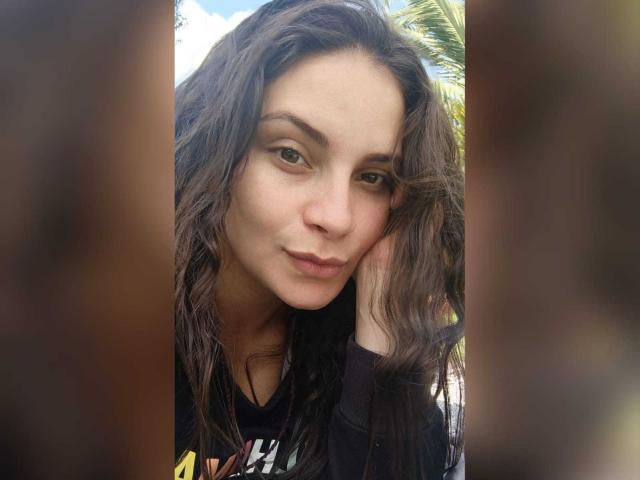 LorelayIncanto has wavy brown hair, light skin, green eyes, and a subtle smile. She's wearing a black shirt, with a hand in her hair, looking relaxed.