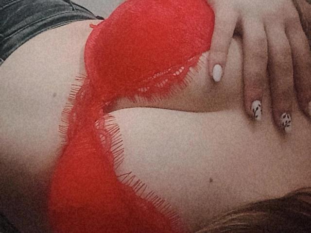 DiLaria's red bra with fringed edges covers her tits, hand with white and patterned nails resting on her chest, skin is pale, and she's wearing black denim.