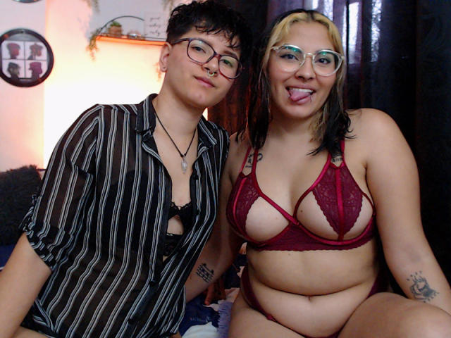 CamilleAndSophie, met kort krullend zwart haar en een bril, draagt een gestreepte shirt en een zwarte beha, zit naast Sophie, met blond en zwart haar, die haar tong uitsteekt, en een rode lingerie aanheeft..