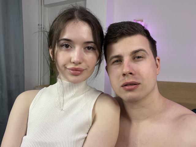 PeterPolly with tits in a white ribbed turtleneck and PeterPolly shirtless, both with fair skin, brown hair, and pinkish lips, standing closely, smiling, in a softly lit room.