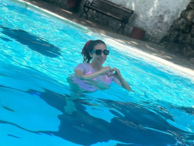 StarBdsm with wet, dark hair in a pink top, wearing black sunglasses, floating in a blue pool, hands near her small, visible tits, water reflecting light.