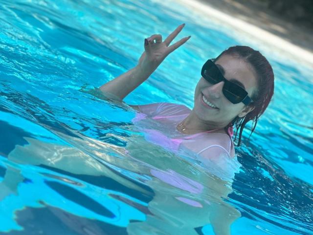 StarBdsm with wet red hair, black sunglasses, and a pink wet shirt, making a peace sign in a blue swimming pool, smiling. Her tits are partially visible.