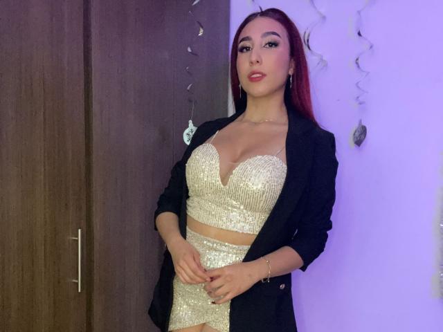 StarBdsm with red hair, wearing a glittery gold crop top and matching skirt, black blazer, and necklace, standing with hands on her belly, showing her tits.