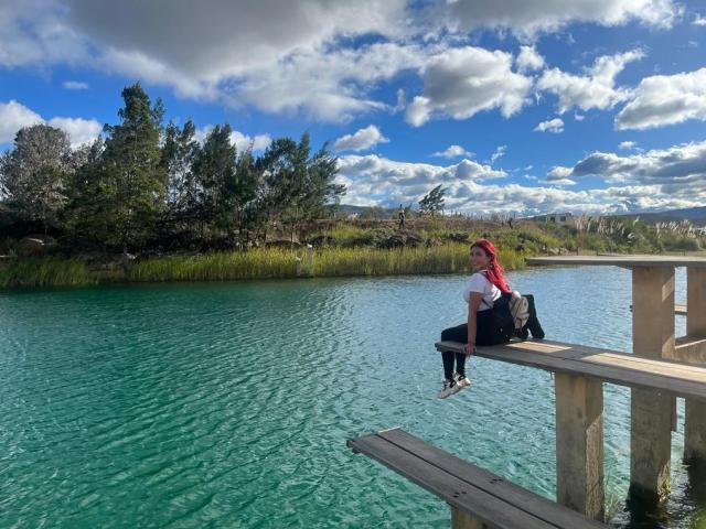 StarBdsm with red hair, white shirt, black pants, and white sneakers, sitting on a wooden dock, looking over a clear green-blue lake, under a partly cloudy sky.