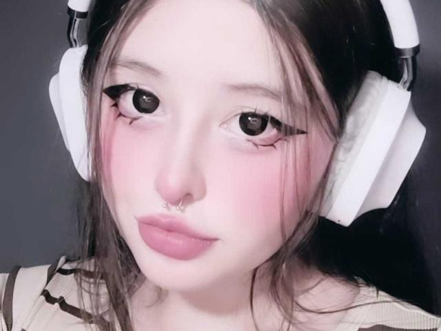 FlutteerShyy has pale skin, pink blush, and pink lips. She wears white headphones, has black eyeliner, and a septum piercing. Her brown hair is slightly messy.