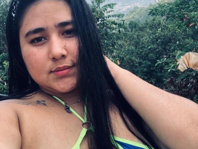ZoeGiil with long black hair, green strappy bikini top, visible tattoo on left shoulder, hand in hair, slight pout, green eyes, looking at camera, outside with greenery.