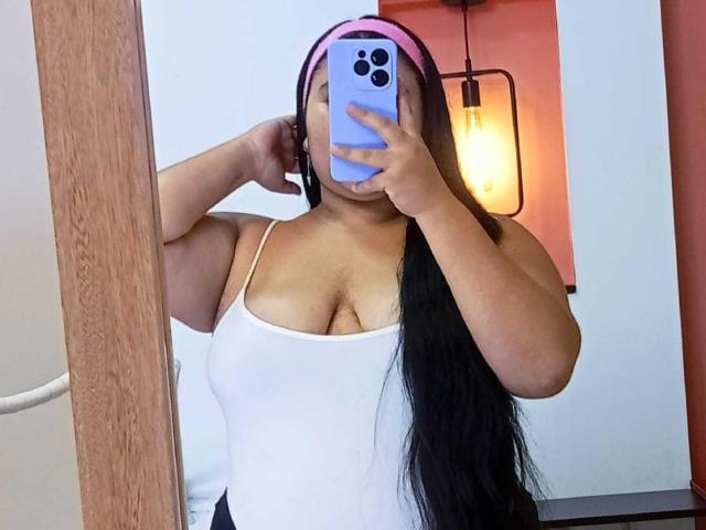 ZoeGiil with long black hair, wearing a white tank top showing her big boobs, pink headphones, holding a blue phone, taking a mirror selfie.