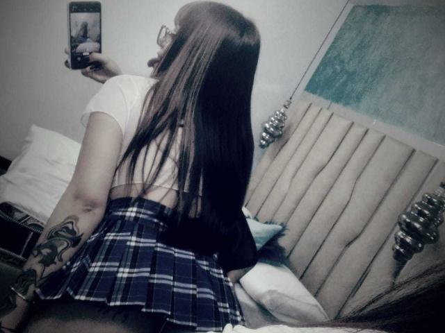 AnniieStone, con cabello castaño largo, usando gafas, una camiseta blanca corta y una falda azul a cuadros, mostrando su brazo tatuado y sentada en una cama, tomando una selfie con un espejo..