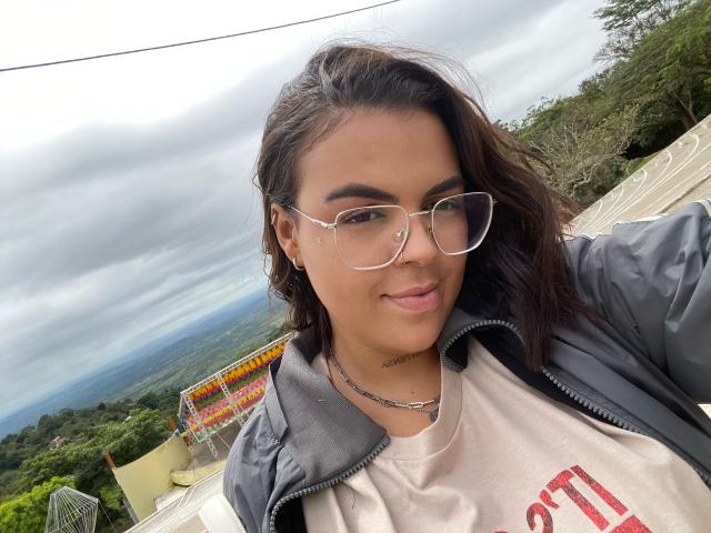 Piettra with glasses, medium brown skin, dark wavy hair, gray jacket, white shirt, silver necklace, smiling, outdoor, cloudy sky, green forest, distant landscape, over-the-shoulder selfie.