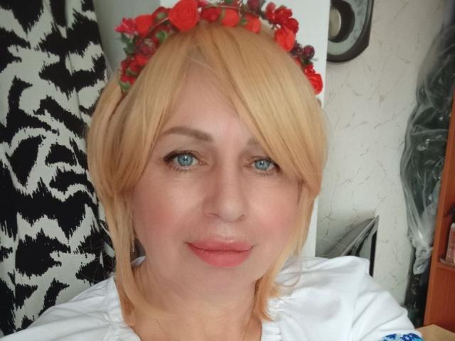 SusieNoreen mit blonden Haaren, blauen Augen und einem roten Blumenkranz, trägt ein weißes Oberteil, hat rosigen Lippenstift und ein leichtes Lächeln. Ihre Brüste sind klein und sie ist süß..