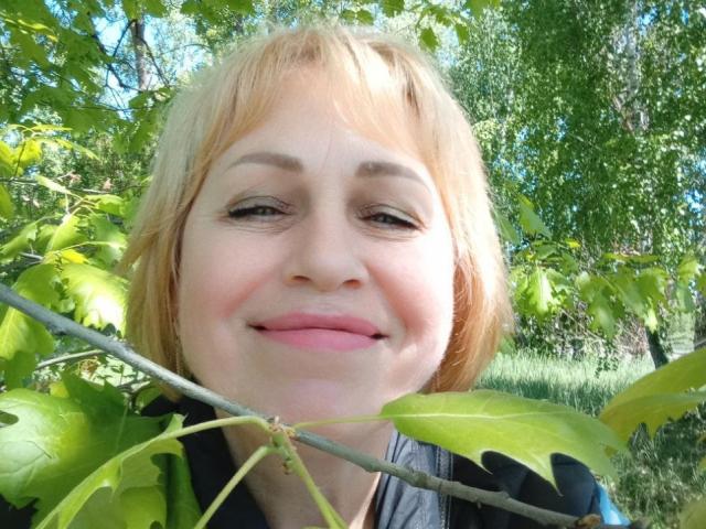 SusieNoreen's face, blonde hair, slightly smiling, green leaves in foreground, blue sky, and trees in background, wearing a black and grey jacket, outdoors.
