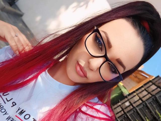 KairaFoxy69, con un cabello que va del rojo al rosa en degradado, gafas negras y una camisa blanca, mostrando su linda cara y un maquillaje sutil. Su mano está apoyada en su hombro..