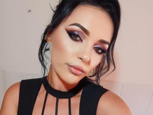 KairaFoxy69 hat dunkles Haar, lila Lidschatten, dicke Augenbrauen, glänzende rosa Lippen und trägt ein schwarzes Oberteil mit Trägern. Sie schaut mit einem leichten Lächeln in die Kamera..