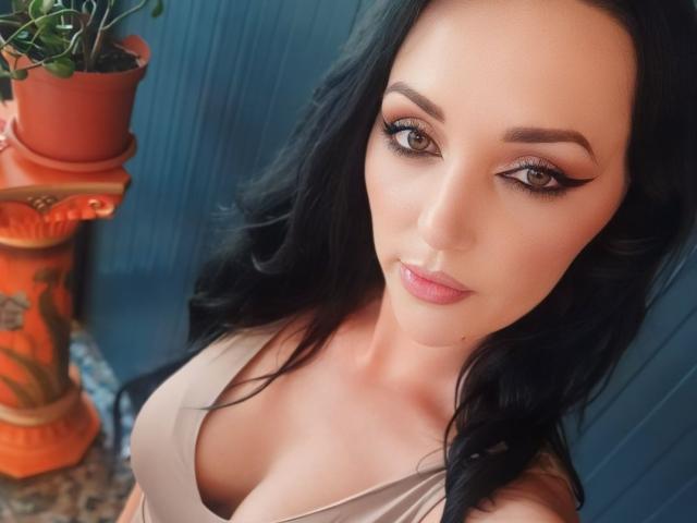 KairaFoxy69 mit schwarzen Haaren, braunen Augen und voller Brüste, trägt ein beiges Oberteil und blickt mit einem leichten Lächeln in die Kamera. Links daneben steht eine Topfpflanze..
