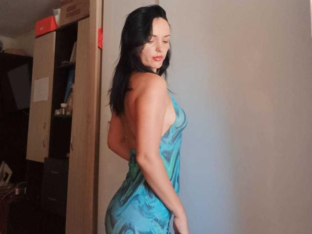 KairaFoxy69 mit schwarzen Haaren, freiem Dekolleté und einem smaragdgrünen Kleid ohne Rücken, das ihren Hintern zeigt, und die über ihre Schulter schaut, in einem schwach beleuchteten Raum..