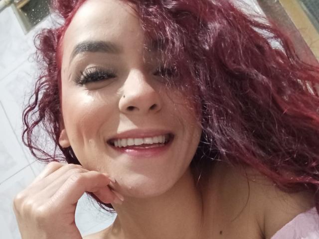 MaiaPark with curly red hair, winking, smiling, nose ring, and hand touching her chin, showing her white teeth and pink lips, with visible breasts in a pink top.