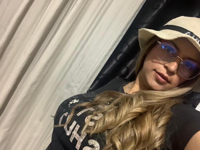 TompsonKarina with long blonde hair, wearing a beige hat, blue glasses, black shirt, and smiling, showing her tits slightly, in a selfie.