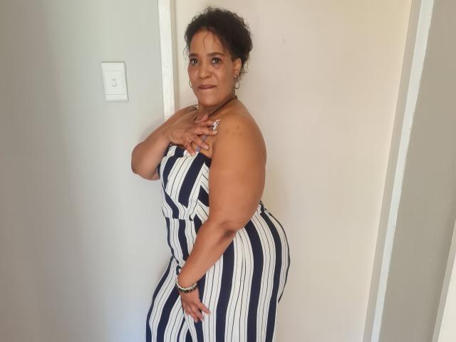 CaramelHugeAss, curvy with big ass, wearing a strapless, navy and white striped dress, standing against a white wall, smiling, dark curly hair, hand on chest, bracelet on wrist.